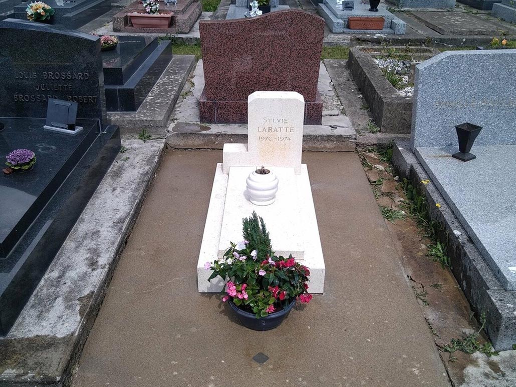 Tombe blanche simple avec gerbe de fleurs et pot de plantes au pied, dans un cimetière, entourée de dalles et de monuments
