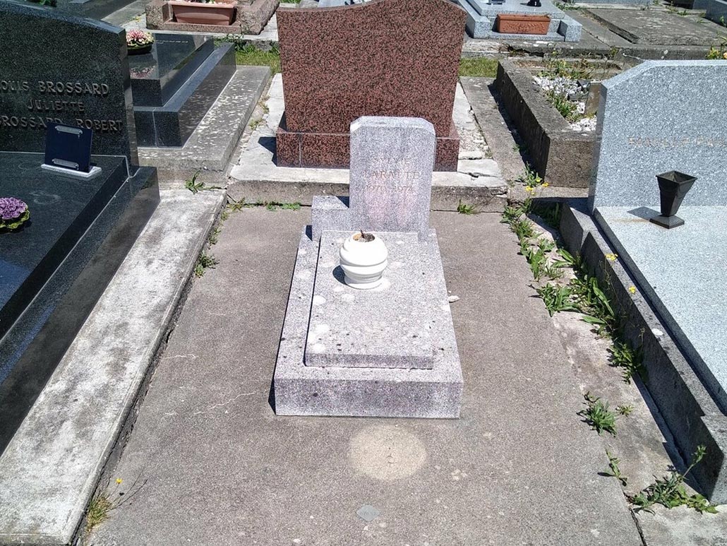 Tombeau en pierre gris clair au milieu d’un cimetière, entouré de monuments funéraires, avec une petite urne blanche sur la