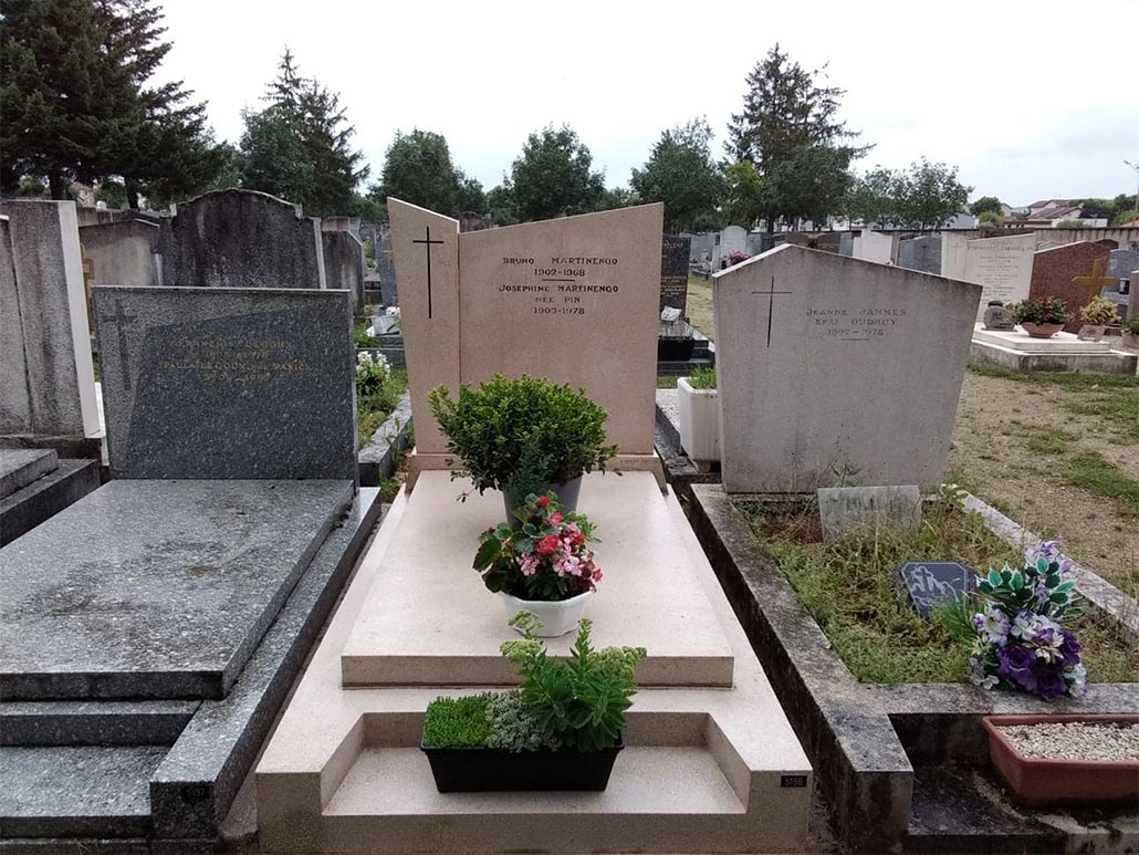 Tombe familiale récemment rénovée dans un cimetière, plaque gravée et fleurs au pied, tombes voisines visibles.