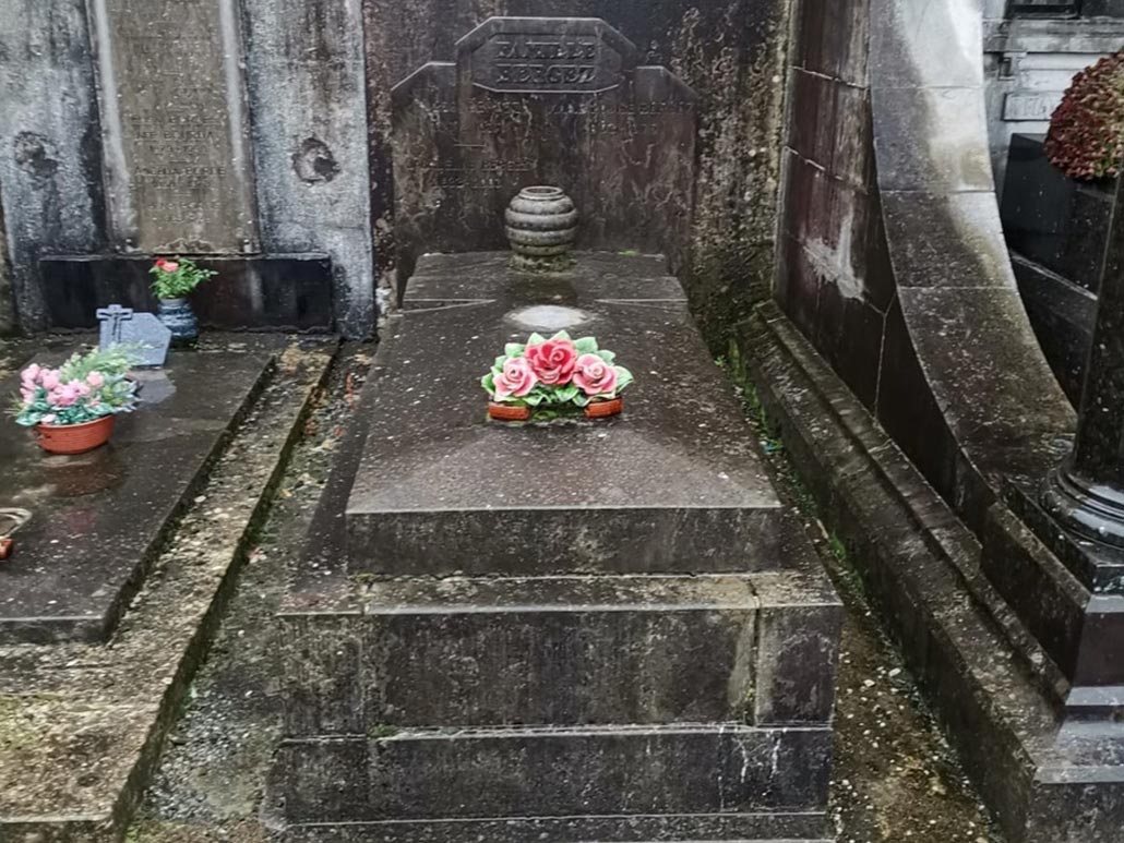 Tombe cimetière en pierre sombre avec bouquet de roses artificielles au centre, entourée de autres monuments funéraires.
