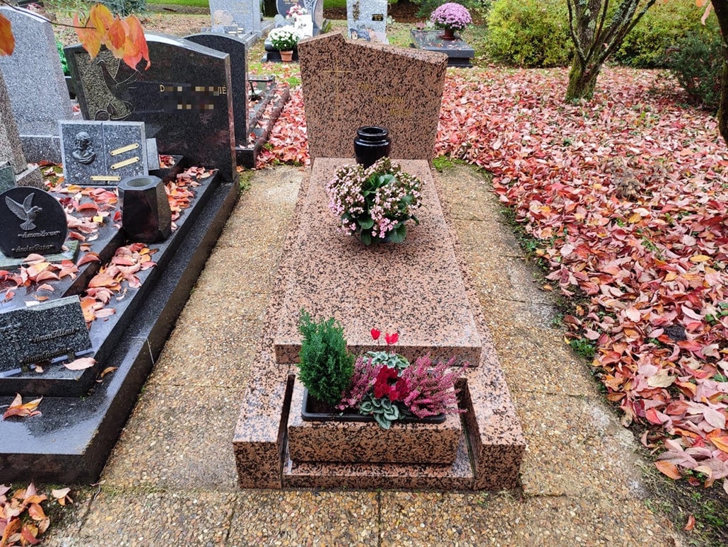 Tombe en pierre rose granit avec jardinière basse contenant des plantes et fleurs, entourée de feuilles d’automne.