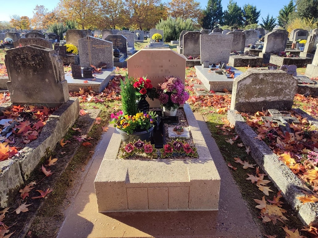 Tombeau paysagé avec fleurs et bouquets colorés sur une stèle en pierre dans un cimetière, feuilles d’automne au sol.