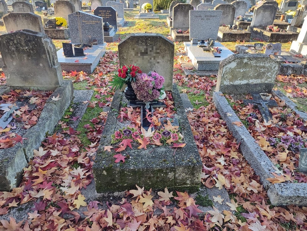 tombe du cimetière spacieux entourée de stèles, feuilles d’automne au sol, gerbes de fleurs colorées déposées sur le tombeau.
