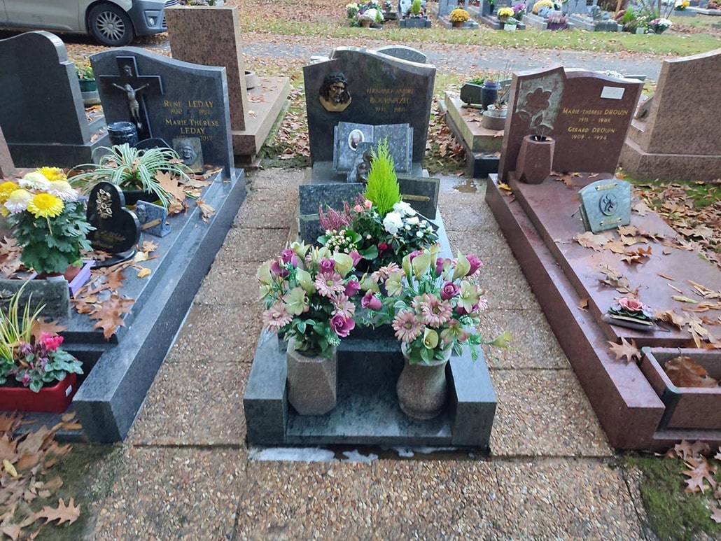 Tombeau gris clair au centre gravé, entouré de bouquets colorés et fleurs artificielles dans des vases; allées couverts de