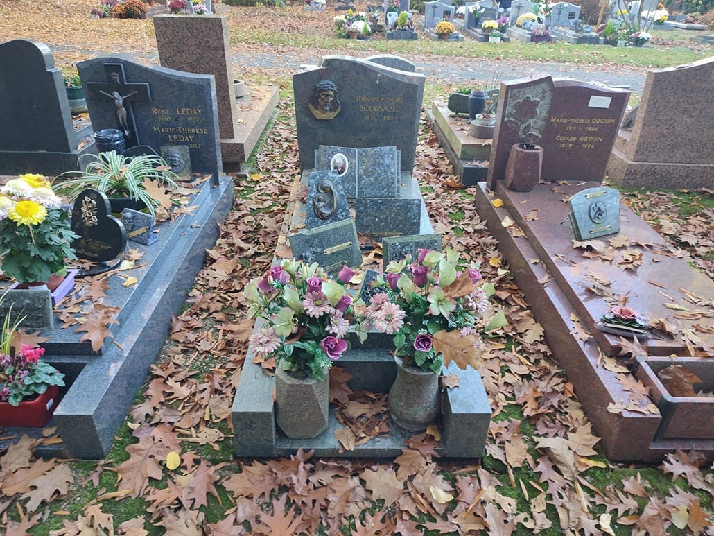 Tombeau familial en pierre grise au cimetière, entouré de feuilles d’automne et de bouquets floraux.