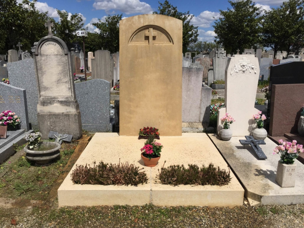 Tombe non répertoriée dans un cimetière, monticule de pierre beige avec croix gravée, fleurs en pot et planteurs.